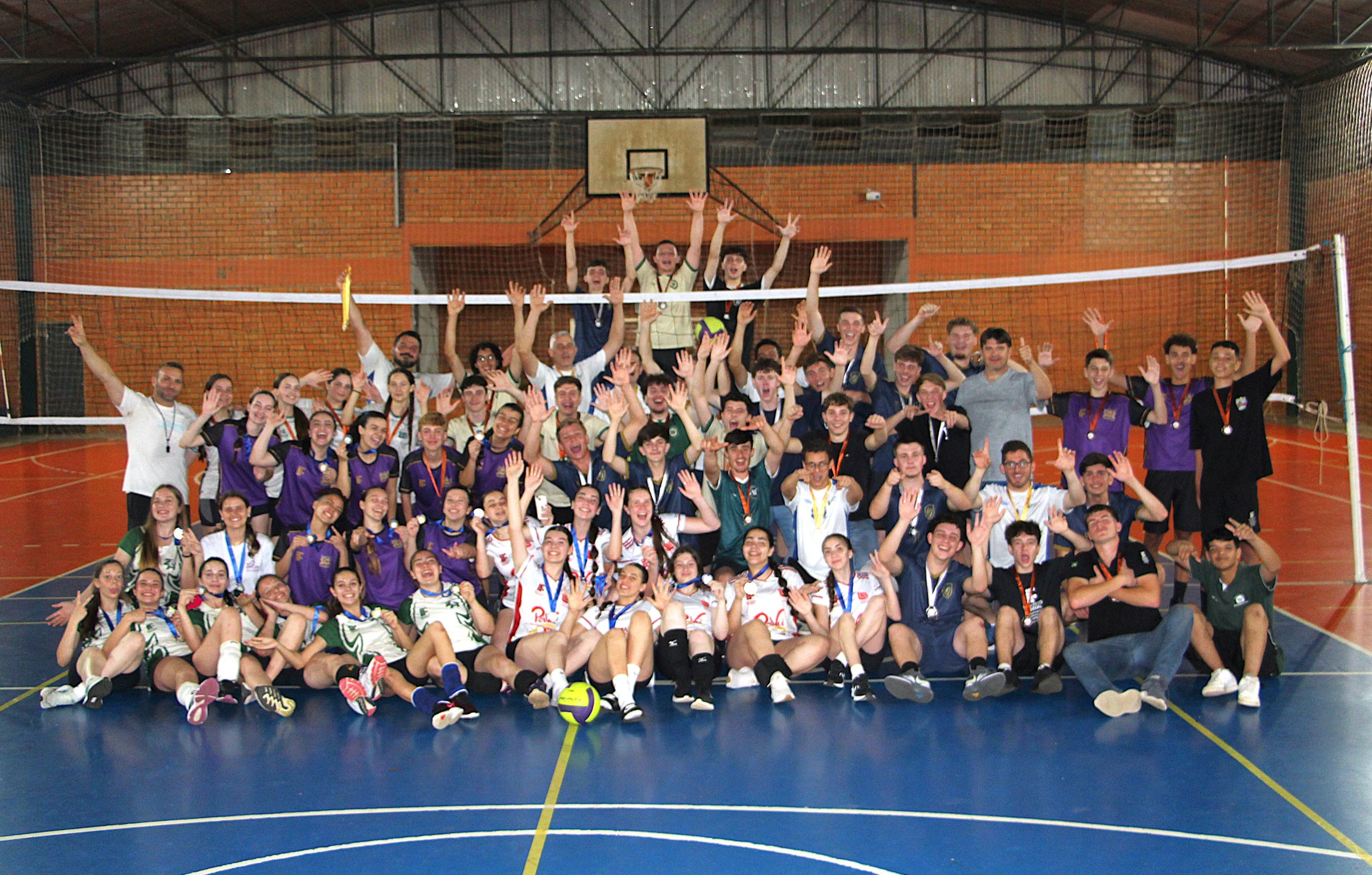 A foto mostra um grupo de aproximadamente 70 pessoas dentro de um ginásio esportivo de tijolinhos aparentes. Elas estão posando para a foto, sentados e em pé sobre uma quadra de piso azul e laranja. Há uma rede de vôlei montada ao fundo do grupo. No grupo há pessoas sorrindo, levantando os braços em comemoração e usando medalhas no pescoço.A maioria está uniformizada para jogos de vôlei.