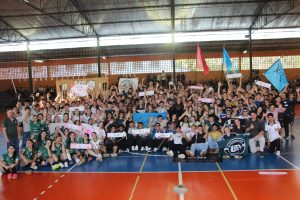 A foto mostra um grupo de aproximadamente 150 pessoas dentro de um ginásio esportivo de tijolinhos aparentes, com uma arquibancada ao fundo. Elas estão posando para a foto, sentados e em pé sobre uma quadra de piso azul e laranja. No grupo há pessoas sorrindo, levantando os braços em comemoração e usando medalhas no pescoço. A maioria está uniformizada para jogos de futsal. Há subgrupos identificados com cartazes e bandeiras em diferentes cores.