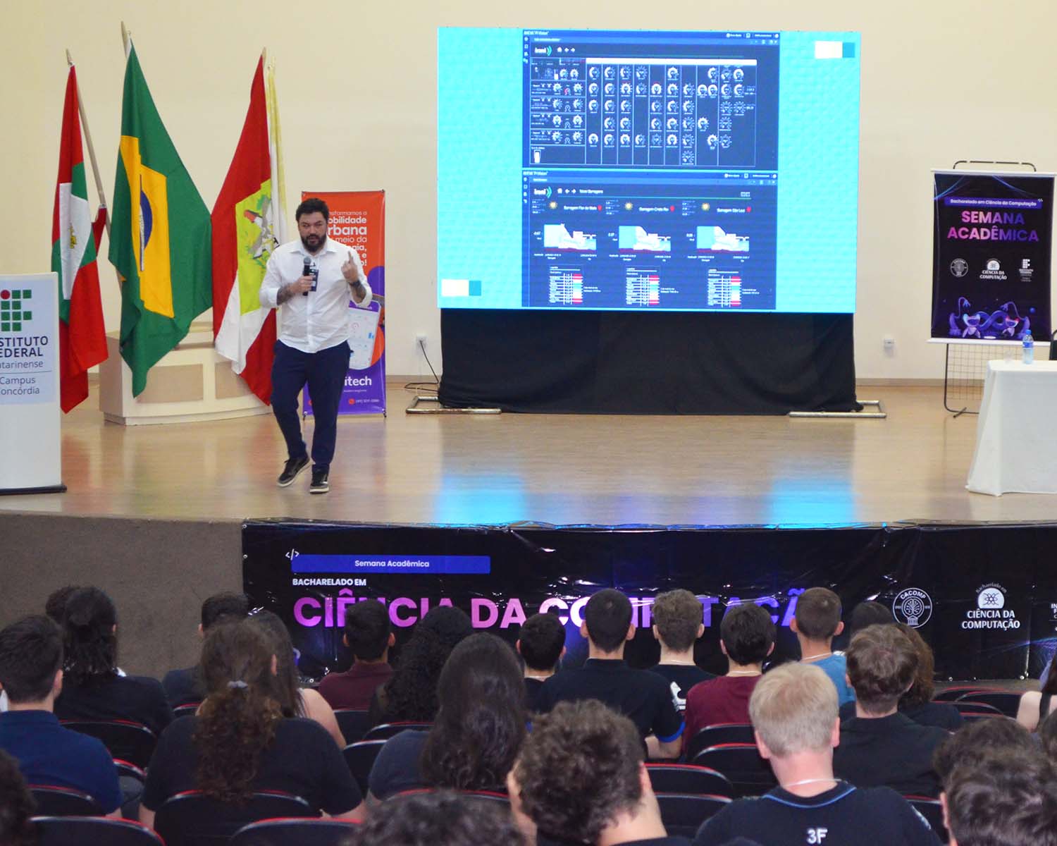 A foto mostra um auditório durante uma palestra. A foto foi tirada dos fundos para a frente, mostrando os alunos assistindo à apresentação. No palco, o palestrante Cristyofer Oliveira veste camisa branca e calça escura; ele segura um microfone e está em movimento, caminhando enquanto fala. À esquerda do palco há um púlpito com o logotipo do IFC Concórdia. Atrás dele estão três bandeiras hasteadas: a de Santa Catarina, a do Brasil e a do município. No centro do palco, um grande telão exibe uma interface gráfica com diversos gráficos e painéis de dados. À direita, um banner com o título “Semana Acadêmica” está exposto em um suporte metálico, e ao lado há uma mesa com uma garrafa de água.