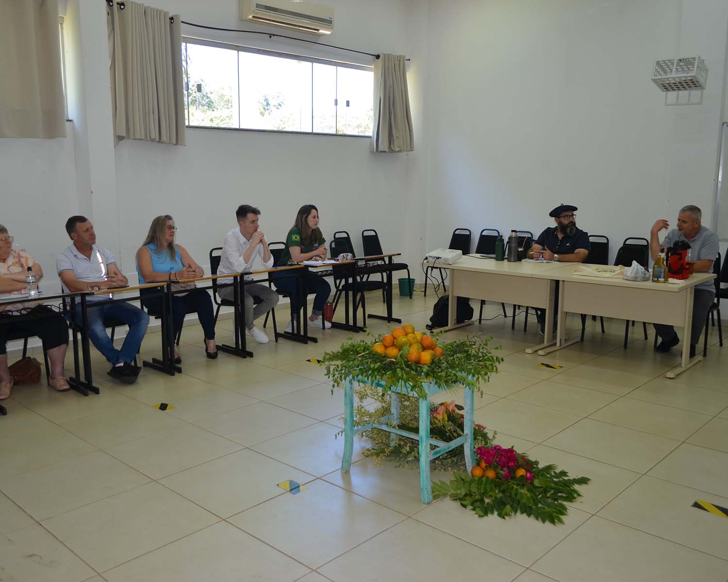 A foto mostra uma sala ampla com paredes brancas e uma janela no alto. Um grupo de pessoas está reunido em formato de semicírculo, sentado em cadeiras atrás de mesas escolares alinhadas lado a lado. Há seis pessoas à esquerda e duas à direita, todas voltadas para o centro da sala, onde está uma pequena mesa azul clara decorada com folhas verdes, flores cor-de-rosa e frutas, principalmente laranjas. Entre os participantes, há homens e mulheres de diferentes idades, vestindo roupas casuais. Um dos homens, à direita, é o professor Edmilson Paglia. Ele fala gesticulando com a mão, enquanto outro, o professor Juliano Schmitz, ao seu lado, o observa atentamente.