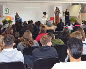 A imagem mostra um auditório com pessoas sentadas, vistas de costas, assistindo a uma mesa-redonda. No palco, há quatro pessoas: uma delas está sentada à esquerda (Luiz Carlos Chaves) , outra está ao centro também sentada com um microfone (Jaqueline Conceição), e duas pessoas estão em pé à direita — uma delas é palestrante (Lumen Lohn Freitas), enquanto a outra realiza tradução em Libras. No fundo, projetado na parede, está o título da fala: “Transfobia e Institucionalidade: Experiência e Resistência em SC”. À esquerda do palco, há um banner comemorativo dos 60 anos do IFC Concórdia, ao lado de uma mesa coberta com tecido branco e uma bandeira da Una LGBT. Na frente, há um arranjo de plantas verdes.