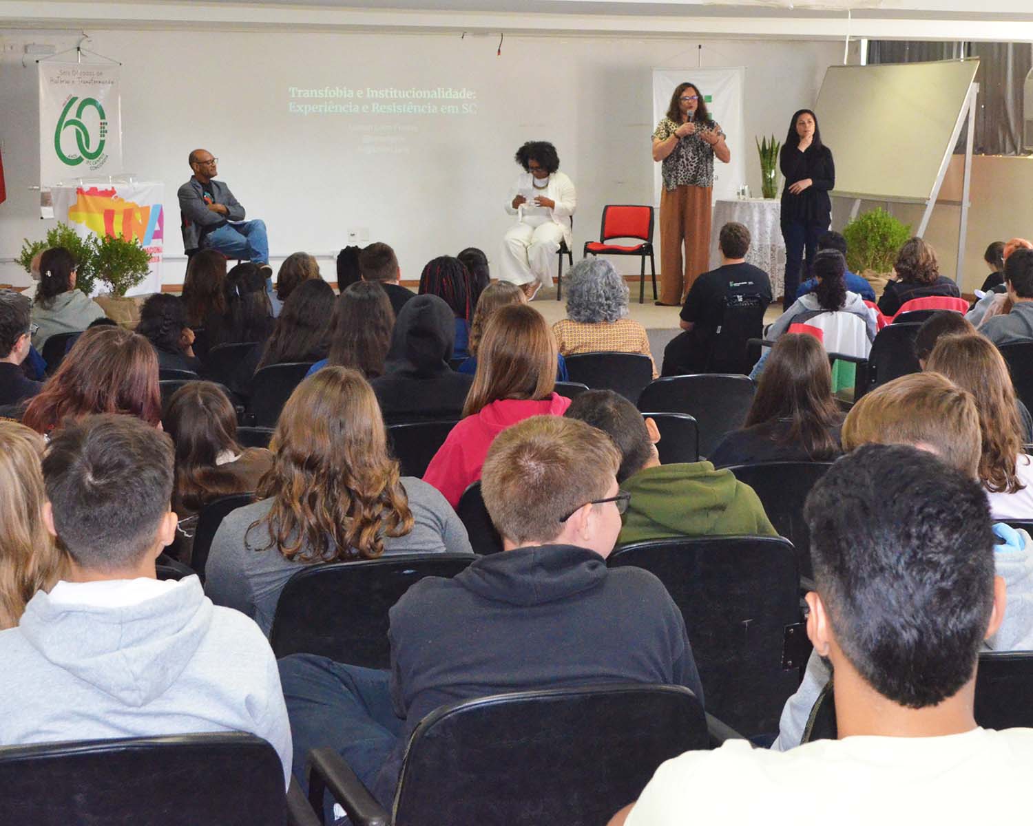 A imagem mostra um auditório com pessoas sentadas, vistas de costas, assistindo a uma mesa-redonda. No palco, há quatro pessoas: uma delas está sentada à esquerda (Luiz Carlos Chaves) , outra está ao centro também sentada com um microfone (Jaqueline Conceição), e duas pessoas estão em pé à direita — uma delas é palestrante (Lumen Lohn Freitas), enquanto a outra realiza tradução em Libras. No fundo, projetado na parede, está o título da fala: “Transfobia e Institucionalidade: Experiência e Resistência em SC”. À esquerda do palco, há um banner comemorativo dos 60 anos do IFC Concórdia, ao lado de uma mesa coberta com tecido branco e uma bandeira da Una LGBT. Na frente, há um arranjo de plantas verdes.