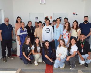 A imagem mostra um grupo de cerca de vinte pessoas posando para uma foto em frente a entrada dos banheiros do bloco pedagógico, um ambiente externo com cobertura e bem iluminado. Elas estão organizadas em duas fileiras: a primeira com jovens sentados agachados, e a segunda com adultos e alguns estudantes em pé. São servidores e estudantes do campus, todos olhando para a câmera. Na parede ao fundo, há duas portas lado a lado, cada uma com um símbolo indicando banheiro feminino e masculino.