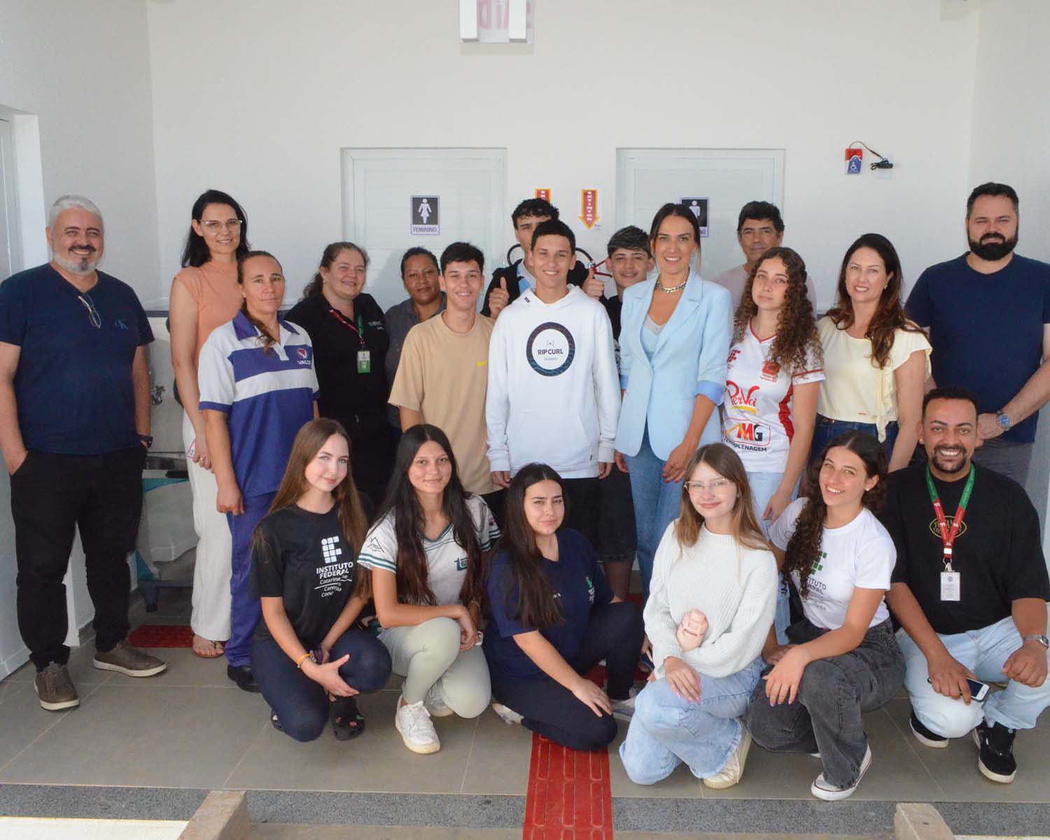 A imagem mostra um grupo de cerca de vinte pessoas posando para uma foto em frente a entrada dos banheiros do bloco pedagógico, um ambiente externo com cobertura e bem iluminado. Elas estão organizadas em duas fileiras: a primeira com jovens sentados agachados, e a segunda com adultos e alguns estudantes em pé. São servidores e estudantes do campus, todos olhando para a câmera. Na parede ao fundo, há duas portas lado a lado, cada uma com um símbolo indicando banheiro feminino e masculino.