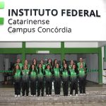 Foto colorida, em ambiente externo, registrada em frente ao prédio administrativo do Instituto Federal Catarinense – Campus Concórdia. Na parte superior da imagem, destaca-se a fachada branca com o logotipo IFC. Em frente à entrada principal do prédio, há um grupo de aproximadamente 18 pessoas, sendo 15 alunas, organizadas em duas fileiras, mais os professores Jerson Isoton e Cleiton Bosio e a técnica adminstrativa Rose Veloso. Todas vestem agasalhos esportivos em tons de verde e preto, com a sigla “IFC”. As pessoas estão de pé, alinhadas, olhando para a câmera e sorrindo.