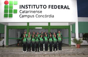 Foto colorida, em ambiente externo, registrada em frente ao prédio administrativo do Instituto Federal Catarinense – Campus Concórdia. Na parte superior da imagem, destaca-se a fachada branca com o logotipo IFC. Em frente à entrada principal do prédio, há um grupo de aproximadamente 18 pessoas, sendo 15 alunas, organizadas em duas fileiras, mais os professores Jerson Isoton e Cleiton Bosio e a técnica adminstrativa Rose Veloso. Todas vestem agasalhos esportivos em tons de verde e preto, com a sigla “IFC”. As pessoas estão de pé, alinhadas, olhando para a câmera e sorrindo.