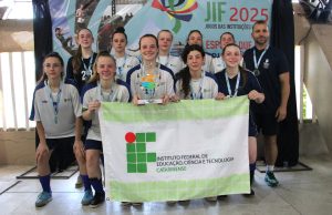 Foto colorida, em ambiente interno.. No centro da imagem, há um grupo de 11 alunas acompanhadas pelo professor Cleiton Bosio à direita. Elas estão organizadas em duas fileiras e vestem uniformes esportivos em tons de branco, azul e verde, com a identificação do Instituto Federal. Todos usam medalhas no peito.