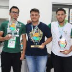 Foto colorida, em ambiente interno. No centro da imagem, há três homens jovens, posicionados lado a lado, olhando para a câmera. Os três usam medalhas penduradas no pescoço e seguram troféus de acrílico transparente com detalhes coloridos e base dourada, relacionados aos Jogos das Instituições Federais – JIF 2025. O homem à esquerda veste camiseta verde com estampa clara e usa óculos de grau. O homem ao centro veste camiseta azul-marinho e jeans, e apoia os braços sobre os ombros dos colegas. O homem à direita veste camiseta branca e verde, com identificação do Instituto Federal.