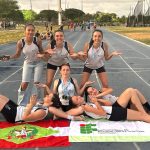Foto colorida, em ambiente externo, registrada em uma pista de atletismo. No centro da imagem, há um grupo de 7 alunas atletas, organizadas de forma descontraída e criativa para a foto. Três estão em pé na parte de trás, com os braços abertos e as palmas das mãos voltadas para cima, sorrindo para a câmera. Uma atleta está sentada ao centro, também sorrindo, usando várias medalhas penduradas no pescoço. Quatro atletas estão deitadas no chão, apoiadas sobre os cotovelos ou reclinadas, formando uma composição simétrica ao redor daquela sentada. Todas vestem uniformes esportivos em tons de branco e preto, com identificação do Instituto Federal. Na parte inferior da imagem, sobre a pista, há duas bandeiras estendidas lado a lado: à esquerda, a bandeira do estado de Santa Catarina; à direita, a bandeira do IFC.
