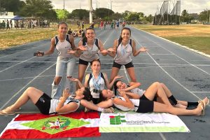 Foto colorida, em ambiente externo, registrada em uma pista de atletismo. No centro da imagem, há um grupo de 7 alunas atletas, organizadas de forma descontraída e criativa para a foto. Três estão em pé na parte de trás, com os braços abertos e as palmas das mãos voltadas para cima, sorrindo para a câmera. Uma atleta está sentada ao centro, também sorrindo, usando várias medalhas penduradas no pescoço. Quatro atletas estão deitadas no chão, apoiadas sobre os cotovelos ou reclinadas, formando uma composição simétrica ao redor daquela sentada. Todas vestem uniformes esportivos em tons de branco e preto, com identificação do Instituto Federal. Na parte inferior da imagem, sobre a pista, há duas bandeiras estendidas lado a lado: à esquerda, a bandeira do estado de Santa Catarina; à direita, a bandeira do IFC.