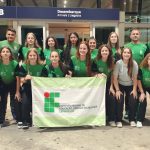 Foto colorida, em ambiente interno, registrada em um aeroporto. No centro da imagem, há um grupo grande de aproximadamente vinte pessoas, entre jovens e adultos, organizadas em duas fileiras. A maioria veste uniformes esportivos em tons de verde e preto, com a sigla IFC estampada nas camisetas e agasalhos. Na fileira da frente, algumas pessoas estão levemente agachadas, enquanto as demais permanecem em pé atrás. No centro do grupo, duas pessoas seguram uma bandeira branca e verde com o logotipo do IFC. Todas as pessoas olham para a câmera, a maioria sorrindo.