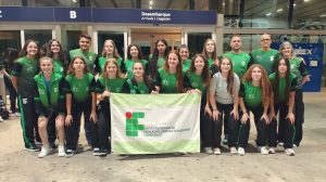 Foto colorida, em ambiente interno, registrada em um aeroporto. No centro da imagem, há um grupo grande de aproximadamente vinte pessoas, entre jovens e adultos, organizadas em duas fileiras. A maioria veste uniformes esportivos em tons de verde e preto, com a sigla IFC estampada nas camisetas e agasalhos. Na fileira da frente, algumas pessoas estão levemente agachadas, enquanto as demais permanecem em pé atrás. No centro do grupo, duas pessoas seguram uma bandeira branca e verde com o logotipo do IFC. Todas as pessoas olham para a câmera, a maioria sorrindo.