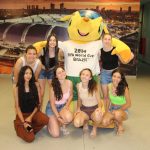 Foto colorida, em ambiente interno. No centro da imagem, há um grupo de seis pessoas, sendo quatro alunas sentadas e duas em pé. Ao lado delas está o mascote da Copa do Mundo do Brasil - 2014.Elas vestem roupas casuais de verão.