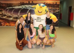 Foto colorida, em ambiente interno. No centro da imagem, há um grupo de seis pessoas, sendo quatro alunas sentadas e duas em pé. Ao lado delas está o mascote da Copa do Mundo do Brasil - 2014.Elas vestem roupas casuais de verão.