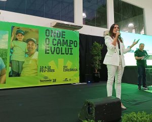 A foto mostra um palco interno com um painel ao fundo em que há uma foto de um homem adulto e uma criança sorrindo, do lado esquerdo, e os dizeres “onde o campo evolui” em letras grandes e “10 a 12 de fevereiro 20206” e “IFC Concórdia” do lado direito, além da logomarcas do evento Tecnoeste, cooperativa Copérdia e Instituto Federal Catarinense no rodapé. Em frente ao painel, há uma mulher (nome: Alessandra Portolan) em pé ao centro e em primeiro plano. Ela veste um conjunto social de blazer e calça branca e sapatos claros, e está falando ao microfone e gesticulando com a mão esquerda.