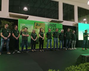 A foto mostra um palco interno com um painel ao fundo em que há uma foto de um homem adulto e uma criança sorrindo, do lado esquerdo, e os dizeres “onde o campo evolui” em letras grandes e “10 a 12 de fevereiro 20206” e “IFC Concórdia” do lado direito, além da logomarcas do evento Tecnoeste, cooperativa Copérdia e Instituto Federal Catarinense no rodapé. Em frente ao painel há 11 pessoas lado a lado, todas olhando pra frente. Elas vestem camisetas verde-escuras com a logo do Tecnoeste no peito. À direita, um homem segura um microfone e parece falar, enquanto os demais permanecem em posição estática, com as mãos juntas à frente do corpo.