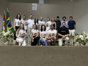 Foto colorida, em ambiente interno, registrada em no palco de um auditório. No centro da imagem, há um grupo de aproximadamente quinze pessoas adultas, mulheres e homens, de diferentes idades e características físicas, organizadas em duas fileiras sobre um palco. Na fileira da frente, cinco pessoas estão ajoelhadas ou sentadas, enquanto as demais permanecem em pé atrás delas. A maioria veste camiseta branca com uma estampa central em verde, vermelho e preto, com o número “15” em destaque - alusivo aos 15 anos do curso. Ao fundo, à esquerda, estão duas bandeiras hasteadas: a do Brasil e a de Santa Catarina. Em ambos os lados do palco, há arranjos florais brancos e verdes em vasos metálicos. As pessoas sorriem e olham em direção à câmera.