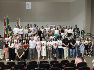 Foto colorida, em ambiente interno, registrada em no palco de um auditório. No centro da imagem há aproximadamente cinquenta pessoas organizadas em várias fileiras. A maioria está em pé; algumas pessoas estão agachadas ou sentadas na parte frontal.
Grande parte do grupo veste camisetas brancas com a estampa do número “15”, enquanto outras pessoas usam roupas variadas, em cores claras e escuras. Há diversidade de gêneros, idades, tons de pele e estilos. Ao fundo, à esquerda, estão duas bandeiras hasteadas: a do Brasil e a de Santa Catarina. Em ambos os lados do palco, há arranjos florais brancos e verdes em vasos metálicos. As pessoas sorriem e olham em direção à câmera.