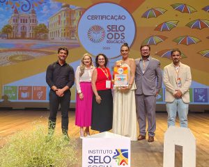 Seis pessoas adultas posam em pé sobre um palco, voltadas para a câmera. Ao centro, uma mulher (Alessandra Portolan) segura uma placa de certificação. Ao lado direito dela está Suzana Scortegagna, e à esquerda está Diego Rodolfo Simões de Lima. O grupo veste roupas formais. Ao fundo, um telão colorido exibe o texto “Certificação Selo ODS Educação 2025”, com elementos gráficos e ilustrações urbanas. No palco, há um arranjo de plantas e um cubo com a inscrição “Instituto Selo Social”.