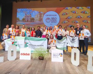 Grupo grande de pessoas adultas reunidas em um palco, posando para foto coletiva. Várias seguram certificados e algumas exibem bandeiras do Instituto Federal Catarinense. À frente, há cubos com logotipos, incluindo “Instituto Selo Social” e “edebê”. O fundo mostra um telão colorido exibe o texto “Certificação Selo ODS Educação 2025”, com elementos gráficos e ilustrações urbanas. O ambiente é um auditório com iluminação voltada para o palco.