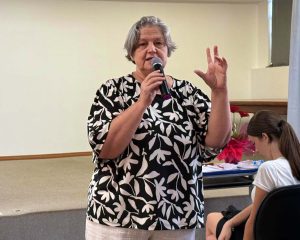 Imagem colorida em ambiente interno. Uma mulher (Dirlei Klein) aparece em primeiro plano. Ela tem pele clara e cabelos curtos grisalhos, veste uma blusa preta com estampa de folhas brancas. Ela segura um microfone próximo ao rosto com a mão direita, enquanto gesticula com a mão esquerda elevada em sinal de explicação. Ao fundo, vê-se uma parede de tom neutro e, no canto inferior direito, parte de um arranjo de flores vermelhas e o perfil de uma jovem sentada.