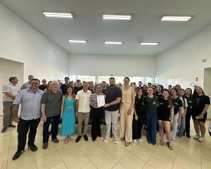 Imagem colorida. Grupo de aproximadamente 50 pessoas posa em pé no hall do prédio administrativo do IFC Concórdia, um salão amplo e bem iluminado, com paredes brancas e luzes no teto. Ao centro, sete adultos estão à frente, sendo que uma mulher segura um documento ao lado de um homem. Ao redor, estudantes e outros participantes se distribuem em semicírculo, olhando para a câmera. O piso é claro e há janelas ao fundo permitindo entrada de luz natural.