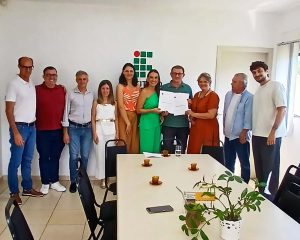 Imagem colorida. Grupo de dez adultos posa em pé em uma sala clara, em frente a uma parede branca com o logotipo do Instituto Federal - a sala de reuniões do prédio administrativo do IFC Concórdia. Ao centro, duas mulheres e um homem seguram um documento aberto, exibido para a câmera. À frente do grupo, há uma mesa retangular com cadeiras ao redor, contendo xícaras de café e um vaso com planta. À direita, uma porta e uma janela deixam entrar luz natural.