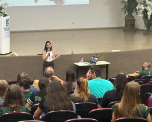 Foto colorida em auditório. Em primeiro plano, pessoas sentadas em poltronas a uma apresentação. À frente da plateia, uma mulher (Adriana Lugaresi) está em pé segurando um microfone. Ela usa blusa branca sem mangas e calça escura e gesticula enquanto fala. Ao lado da palestrante, há uma mesa com um notebook aberto, uma garrafa de água e um copo. Ao fundo, há uma tela de projeção. À esquerda do palco, há um púlpito com o logotipo do Instituto Federal Catarinense – Campus Concórdia. À direita, há um arranjo de flores grandes.