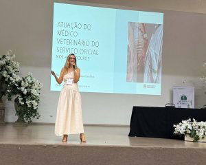 Mulher em pé sobre um palco segura um microfone e apresenta um slide projetado ao fundo. Ela veste vestido claro e usa crachá. O slide traz o título “Atuação do médico veterinário do serviço oficial nos abatedouros”, acompanhado de uma imagem lateral. No palco, há arranjos de flores brancas e uma mesa com toalha preta.
