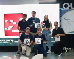 Foto colorida. Seis jovens posam em frente a um painel do “Hackathon do Comércio”. Três estão em pé e três ajoelhados. Todos seguram troféus ou caixas de fones de ouvido recebidos como premiação.