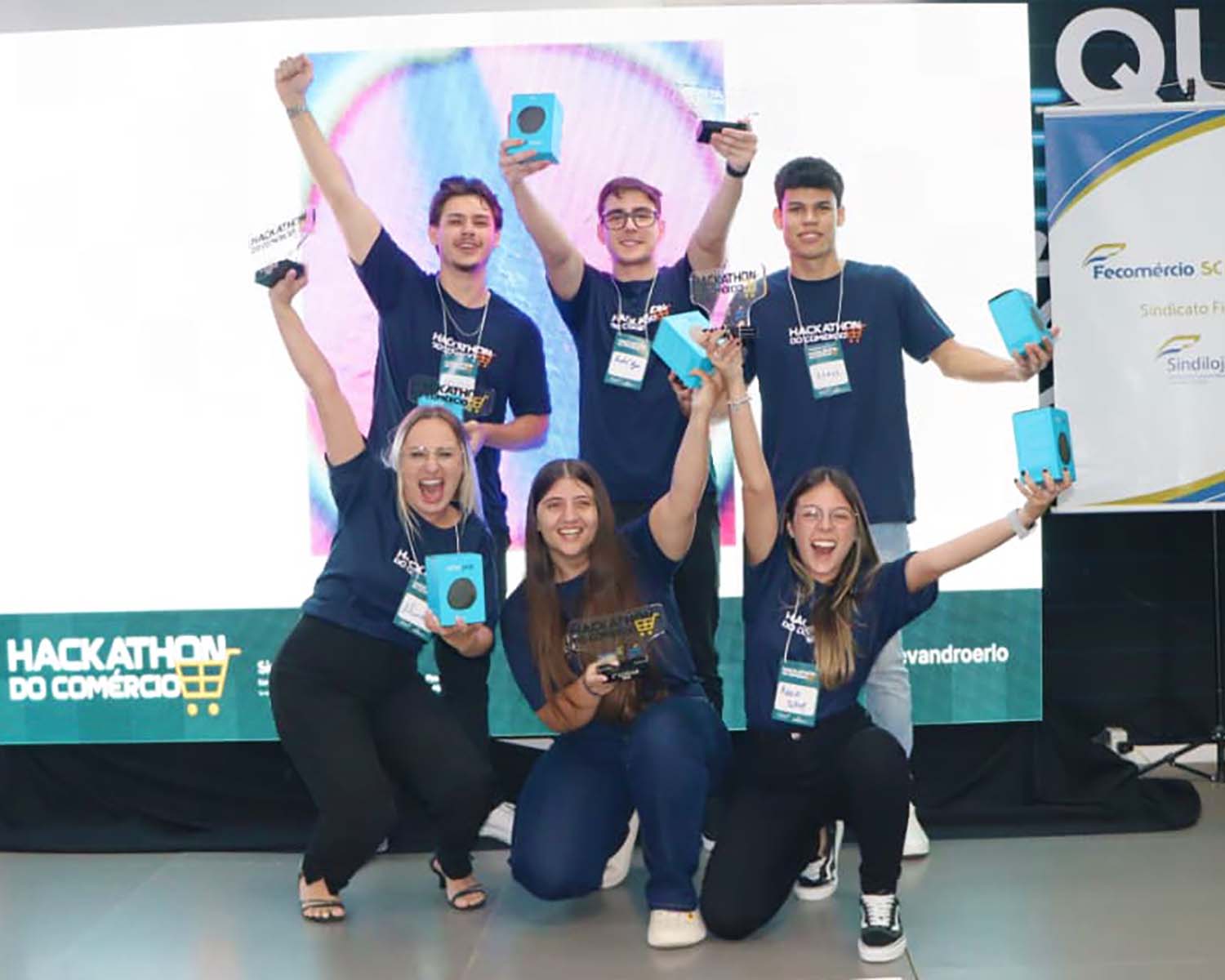 Seis jovens comemoram a premiação do hackathon. Três estão em pé ao fundo e três ajoelhados à frente, com os braços erguidos, segurando troféus e caixas azuis.