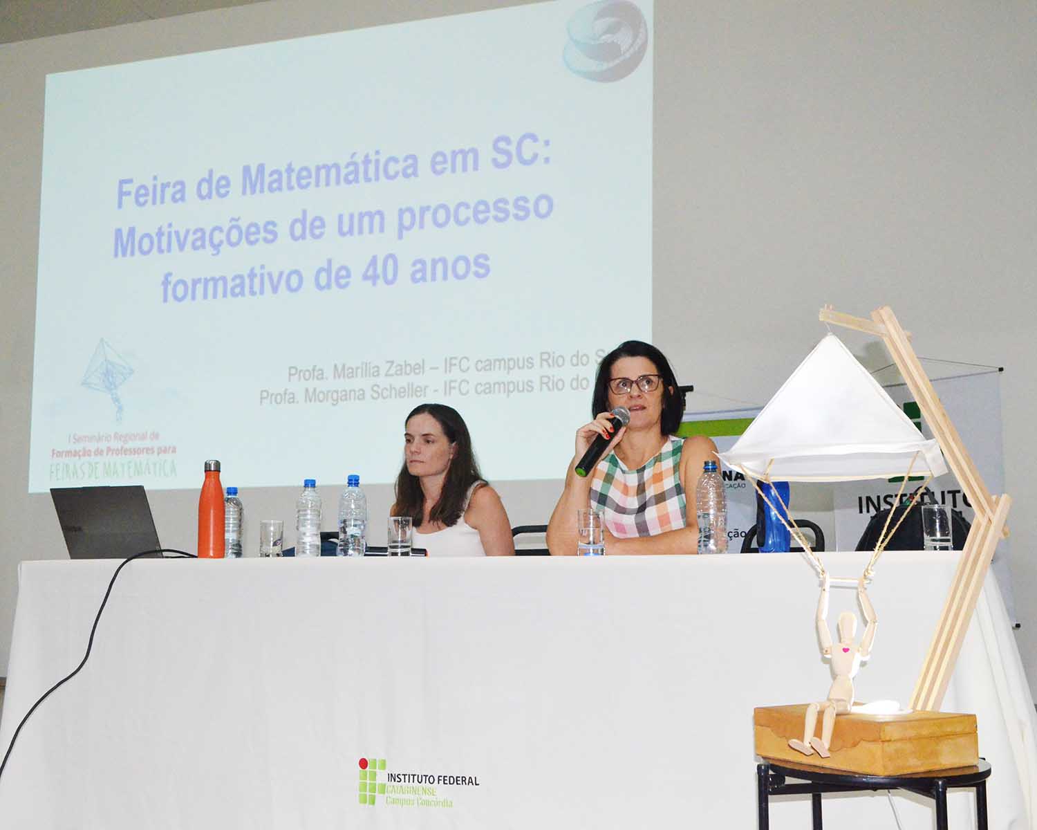 Duas mulheres estão sentadas atrás de uma mesa em um auditório, durante uma apresentação. A mulher à direita segura um microfone e fala, enquanto a outra observa. Sobre a mesa há garrafas de água, copos e um notebook. Ao fundo, um telão projeta o título: “Feira de Matemática em SC: Motivações de um processo formativo de 40 anos”, com nomes de professoras e identificação do IFC. À direita da mesa, há um objeto decorativo de madeira com uma pequena figura suspensa remetendo ao paraquedas de Da Vinci.