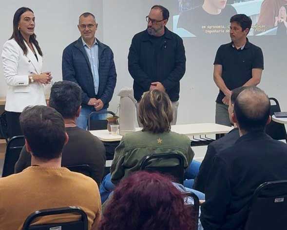 Sala de reunião com público sentado em cadeiras, visto de costas. À frente, quatro pessoas estão em pé: uma mulher à esquerda fala, enquanto três homens ao lado observam. Há uma mesa com jarra térmica e copos. Ao fundo, uma projeção em tela com imagem parcialmente visível. O ambiente é interno, com paredes claras e disposição informal para apresentação.