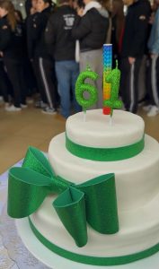 Imagem de um bolo branco de dois andares decorado com detalhes em verde brilhante. Na frente do bolo há um grande laço verde. No topo, velas em formato do número “61”, além de uma vela colorida ao centro. Ao fundo, desfocado, aparece um grupo de pessoas em pé.