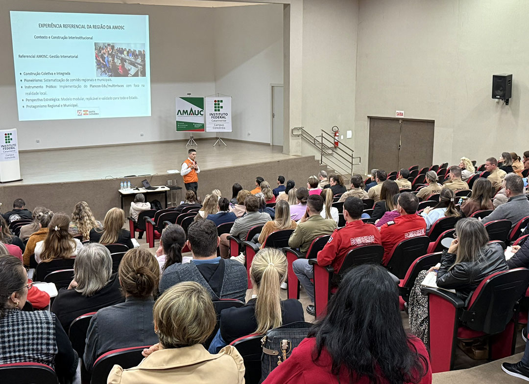 Imagem de um auditório com grande público sentado em cadeiras vermelhas, acompanhando uma palestra. No palco, um homem com uniforme laranja fala ao público segurando um microfone. Ao fundo, um telão exibe uma apresentação segurança em escolas. Próximo ao palco, aparecem banners da AMAUC e do Instituto Federal Catarinense – Campus Concórdia. Entre os participantes, há pessoas com uniformes da Defesa Civil, bombeiros e outras instituições.