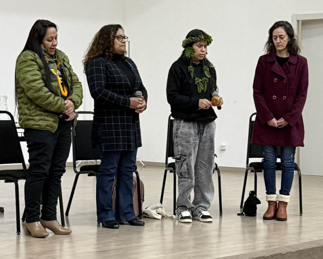 Quatro pessoas estão em pé sobre um palco, em um ambiente interno de paredes claras - o Auditório Central. Da esquerda para a direita: uma mulher com casaco verde, calça preta e botas beges; uma mulher de cabelos cacheados, usando casaco xadrez azul-marinho e óculos; uma mulher com adornos indígenas em tons de verde na cabeça e no pescoço, moletom preto e calça cinza, segurando um maracá; e uma mulher com casaco vinho, jeans e botas marrons. Algumas cadeiras pretas aparecem ao fundo.