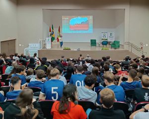 Auditório amplo com público numeroso sentado em cadeiras voltadas para o palco. A plateia é composta majoritariamente por jovens. No palco, uma mulher apresenta em pé, ao centro, segurando um microfone. Ao fundo, há uma projeção com slide sobre águas subterrâneas, com gráfico e texto. À esquerda do palco, estão as bandeiras do Brasil e de Santa Catarina e um púlpito com logotipo institucional. À direita, há duas poltronas verdes e um pequeno espaço montado para conversa. O ambiente é interno, bem iluminado, com foco na apresentação. 