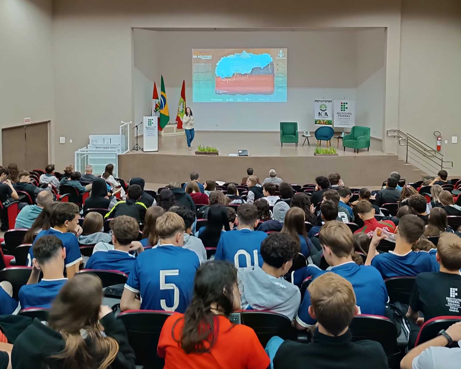 Auditório amplo com público numeroso sentado em cadeiras voltadas para o palco. A plateia é composta majoritariamente por jovens. No palco, uma mulher apresenta em pé, ao centro, segurando um microfone. Ao fundo, há uma projeção com slide sobre águas subterrâneas, com gráfico e texto. À esquerda do palco, estão as bandeiras do Brasil e de Santa Catarina e um púlpito com logotipo institucional. À direita, há duas poltronas verdes e um pequeno espaço montado para conversa. O ambiente é interno, bem iluminado, com foco na apresentação.