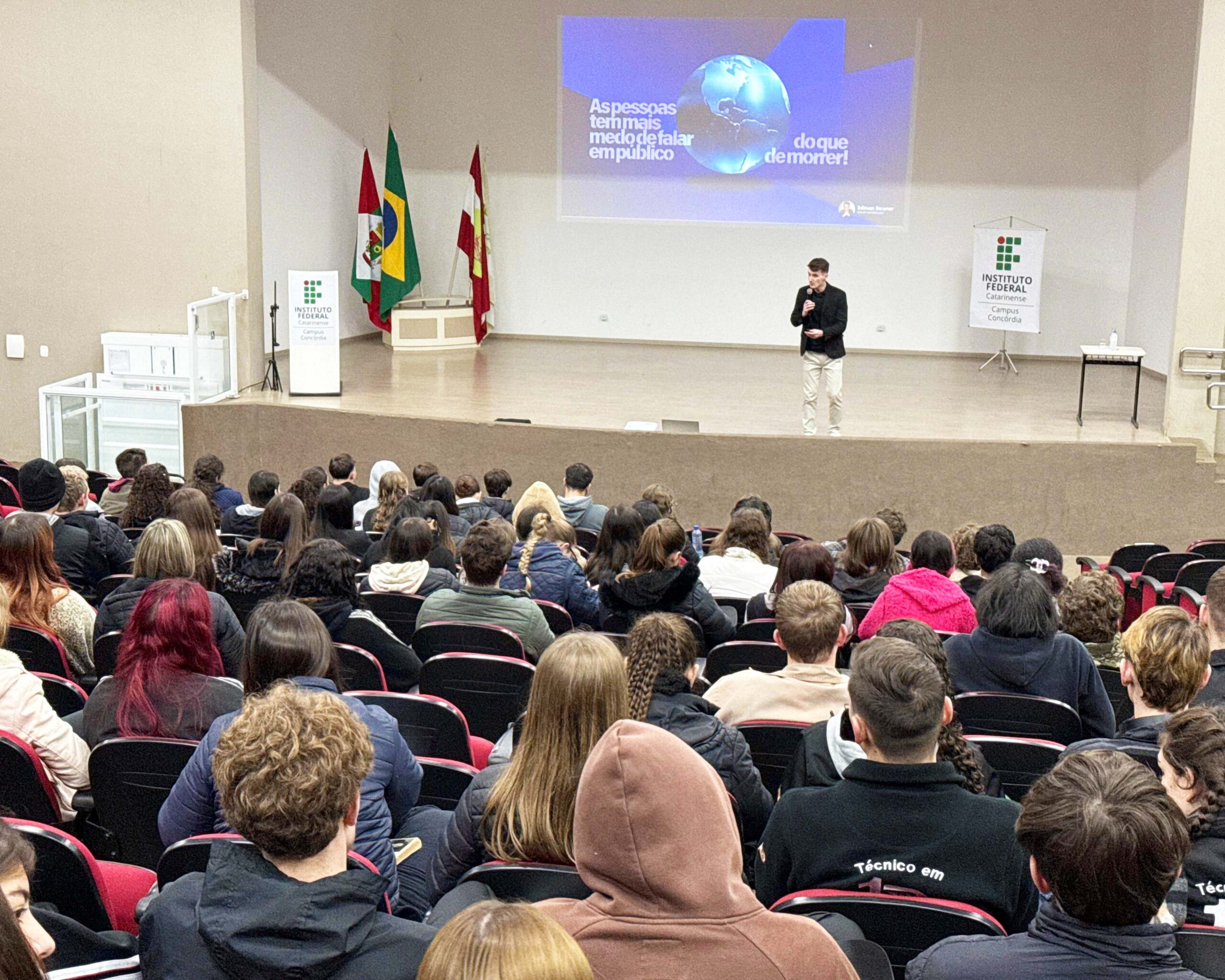 Fotografia de um auditório lotado durante uma palestra. Em primeiro plano, dezenas de pessoas estão sentadas em poltronas vermelhas, voltadas para o palco. No centro do palco, um homem jovem segura um microfone e fala ao público. Ao fundo, uma projeção exibe o slide com a frase: “As pessoas têm mais medo de falar em público do que de morrer!”. Bandeiras do Brasil, de Santa Catarina e do IFC estão posicionadas à esquerda, e banners do Instituto Federal Catarinense Campus Concórdia aparecem nas laterais do palco. O ambiente é amplo, iluminado e de paredes claras.