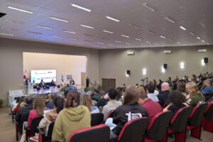Fotografia de um auditório amplo, com dezenas de pessoas sentadas em cadeiras, voltadas para um palco. No palco, há uma mesa com participantes sentados, um púlpito e uma pessoa em pé ao centro. Ao fundo, um telão exibe a identidade visual do IX Seminário da Educação Profissional Técnica Integrada ao Ensino Médio do IFC. Há bandeiras e materiais institucionais nas laterais. A imagem registra um momento de debate durante o evento.