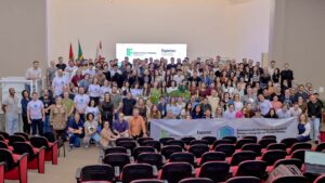 Fotografia colorida em plano geral e angulação superior do palco de um auditório no IFC Campus Concórdia. Dezenas de pessoas de diversas idades estão reunidas no palco em pé e ajoelhadas na frente. No centro da primeira fileira, alguns participantes seguram uma faixa branca horizontal com a inscrição "Seminário da Educação Profissional Técnica Integrada ao Ensino Médio do IFC" e os logotipos do Instituto Federal Catarinense e da FAPESC. A parede ao fundo é branca e conta com uma tela de projeção exibindo os mesmos logotipos e textos da faixa. À esquerda, há mastros com as bandeiras do Brasil, de Santa Catarina e institucionais.